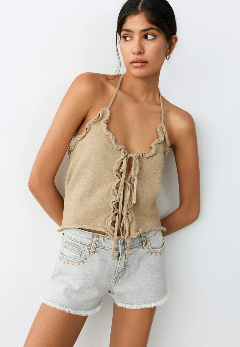 Jeune femme portant un top beige dos-nu avec des volants et un détail noué à l'avant, associé à un short en jean clair clouté, debout devant un fond blanc.