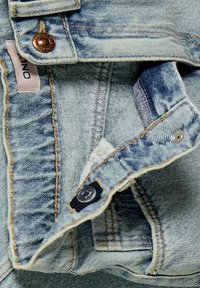 Veste en denim bleu clair avec quincaillerie cuivrée, coutures visibles et un accent de bouton bleu marine. Présente une texture douce et des zones déchirées.