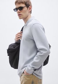 Μακρυμάνικο πόλο σε ανοιχτό γκρι με υφασμάτινη υφή, μαύρο σακίδιο πλάτης και γυαλιά ηλίου. Τζιν παντελόνι ολοκληρώνει το casual ντύσιμο.