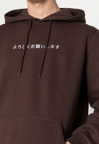 Felpa in cotone marrone con tasca frontale e cappuccio con cordino. Contiene testo bianco in giapponese, che indica un saluto o una richiesta.