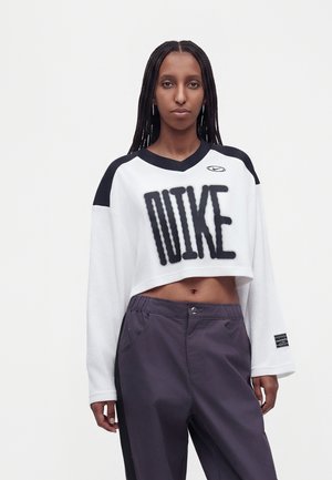 Жена позира с късо бяло дълго ръкаво на Nike с черни букви и тъмни панталони пред едноцветен светъл фон.