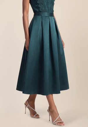 Frau trägt einen taubenblauen plissierten Midirock, ein Spitzentop und silberne Riemchen-Sandaletten mit hohen Absätzen, steht vor einem beigen Hintergrund.