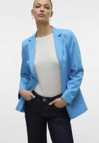 Blazer azzurro chiaro con un design sartoriale, indossato sopra una maglietta bianca e jeans in denim scuro. Presenta rever e tasche laterali. Texture liscia.