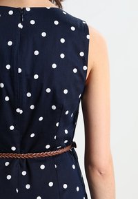 Ärmelloses dunkelblaues Kleid mit weißen Punkten, ausgestattet mit einem geflochtenen braunen Gürtel in der Taille und einem Reißverschlussdetail auf der Rückseite.