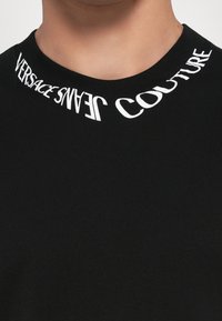 Svart bomull T-skjorte med rund hals. Utringningen viser "VERSACE JEANS COUTURE" i fet hvit skrift, delvis buet.