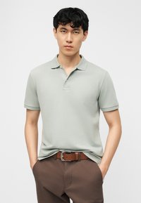 Νέος άνδρας με μαύρα μαλλιά που φοράει ανοιχτοπράσινο πόλο μπλουζάκι και καφέ παντελόνι, στέκεται με τα χέρια στις τσέπες μπροστά σε ενιαίο φόντο.