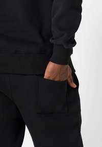 Schwarzer Sweatshirt mit gerippten Bündchen, kombiniert mit schwarzen Hosen, die eine große Tasche haben, in der eine Hand liegt. Weicher, strukturiertes Gewebe.