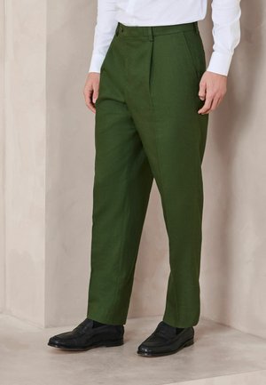 Mand iført skræddersyede olivengrønne bukser, hvid skjorte og sorte læderloafers, stående indendørs mod en lysefarvet væg.