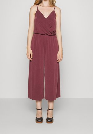 Burgunderfarbener Jumpsuit mit einem verdrehten Vorderdesign, verstellbaren Spaghettiträgern und weiten, gekürzten Beinen. Kombiniert mit schwarzen Plateauschuhen.