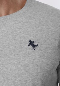 Κοντινή λήψη ενός ατόμου που φοράει ανοιχτό γκρι πουκάμισο με ένα μικρό κεντητό σκούρο έμβλημα ενός ατόμου σε όρθιο άλογο στο στήθος.