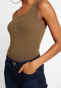 Olivgrön ribbad linne med rund halsringning och tre knappar, tillsammans med mörkblå denimjeans.