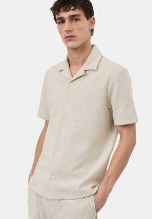 Ung mand iført en beige, tekstureret kortærmet skjorte med knapper og matchende bukser, stående med den ene hånd i lommen mod en hvid baggrund.