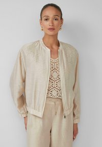 Giacca bomber beige chiaro con una texture liscia, polsini a coste e chiusura a zip, indossata sopra un top all'uncinetto. Design semplice, vestibilità casual.
