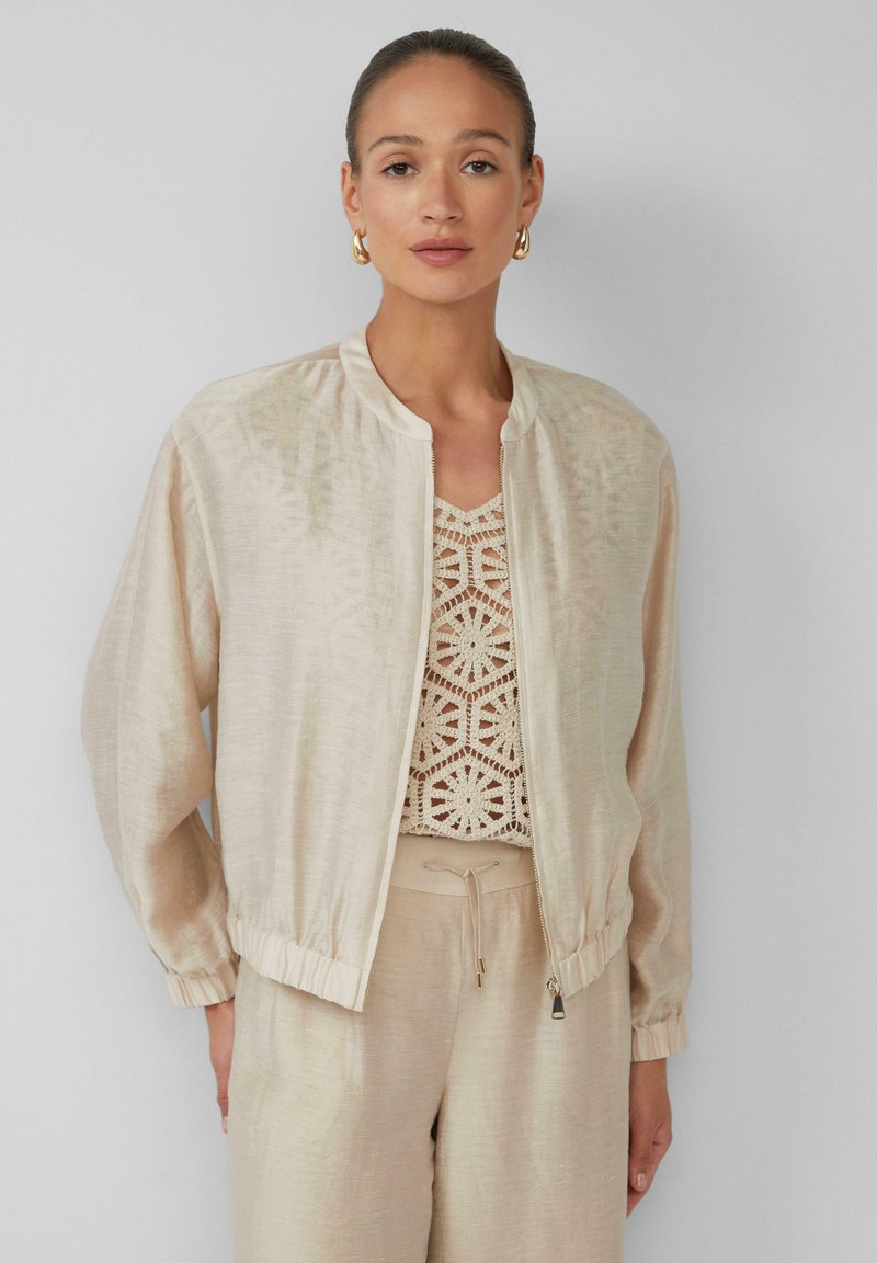 Giacca bomber beige chiaro con una texture liscia, polsini a coste e chiusura a zip, indossata sopra un top all'uncinetto. Design semplice, vestibilità casual.