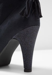 Zapato de tacón alto de ante negro que presenta un tacón negro brillante con un diseño afilado y un acabado texturizado, complementado con detalles de borlas.