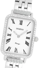 Montre rectangulaire argentée avec un cadran orné de cristaux, un fond blanc, des chiffres romains noirs et un bracelet en maillons métalliques.