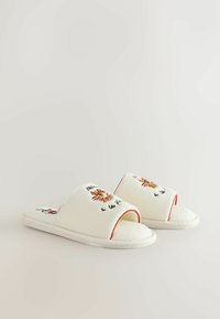 Zapatillas abiertas blancas con sol bordado y texto en la tira superior, y una suela plana, sobre un fondo claro.