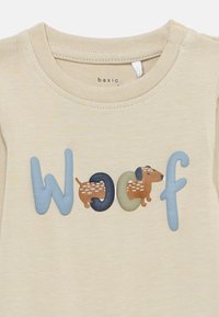 Beige Hemd mit dem Wort "Woof", wobei eine braune Dackelhund-Illustration in die Buchstaben integriert ist.