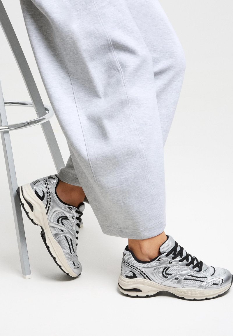 Personne portant des chaussures de sport argentées et noires avec des lacets noirs, un pantalon gris à jambes larges, assise sur un tabouret en métal avec un pied au sol.
