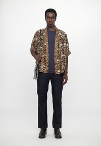 Hnědá a krémová košile s krátkým rukávem a vzorem, širokým límcem, v kombinaci s námořnickým tričkem, tmavými džíny a hnědými botami, držící tašku.