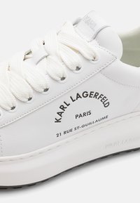 Zapatilla de cuero blanca con suela gruesa, cordones blancos texturizados, branding en texto negro en el lateral y un sutil acento gris en la lengüeta.