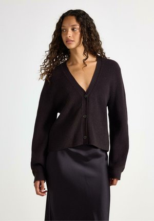 Donna con capelli ricci indossa un cardigan scuro a coste con bottoni sopra una gonna in satin nero su uno sfondo chiaro e uniforme.