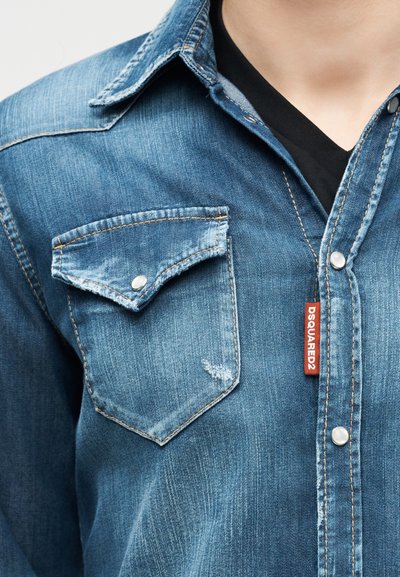 Κοντινό πλάνο ενός ατόμου που φοράει μπλε πουκάμισο από τζιν με τσεπάκι στο στήθος με κουμπιά και κόκκινη ετικέτα "DSQUARED2", πάνω από ένα μαύρο T-shirt.