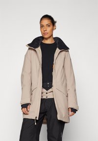 Parka beige con cuello de piel sintética azul marino, cierre frontal con cremallera y múltiples bolsillos. Llevado sobre una camiseta negra de manga larga, combinado con pantalones negros.