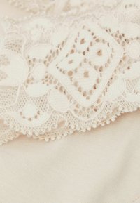 Crème kantstof met ingewikkelde bloemmotieven en golfranden, gelaagd over een glad, effen crème textielachtergrond.