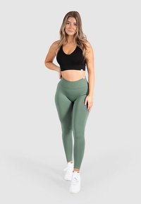 Top corto negro y leggings verdes de cintura alta, hechos de material elástico, combinados con zapatillas blancas, posando sobre un fondo gris claro.
