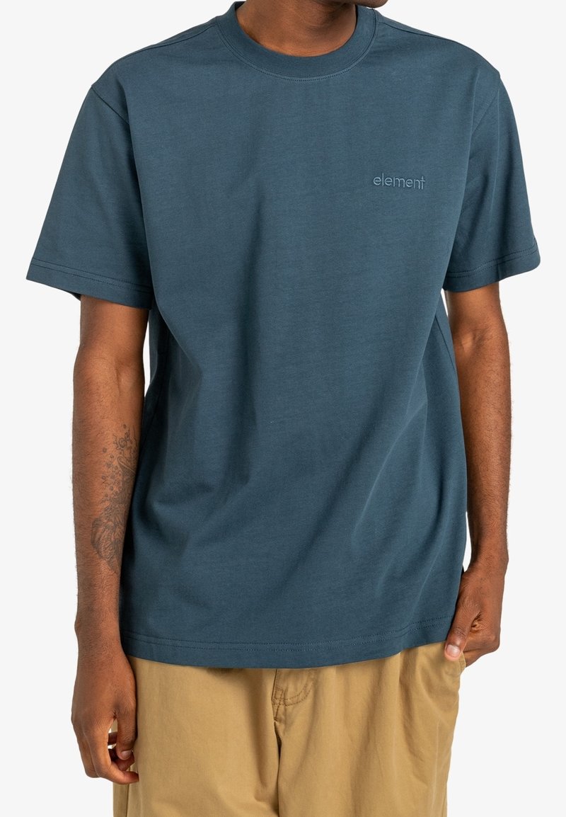 Homme portant un t-shirt à manches courtes uni bleu sarcelle foncé avec le logo "element" sur la poitrine et un pantalon marron clair, debout les mains détendues.