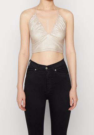 Kvinde iført en metalklar beige halter crop top og højtaljede sorte skinny jeans, stående mod en ensfarvet baggrund.