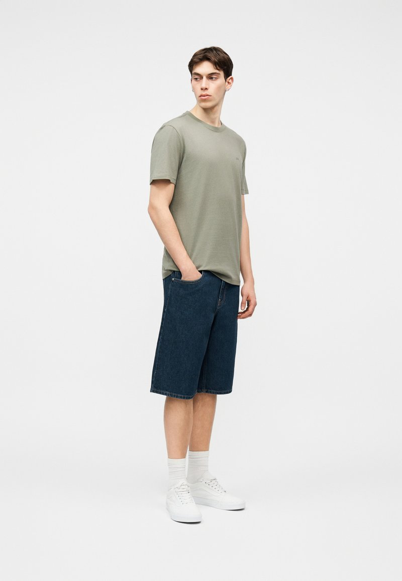 Νεαρός άνδρας που φοράει ανοιχτοπράσινη κοντομάνικη μπλούζα, σκούρα τζιν σορτς μέχρι τα γόνατα, λευκές κάλτσες και λευκά αθλητικά παπούτσια, στέκεται με το ένα χέρι στην τσέπη.