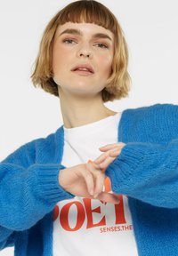 Blauer Strickcardigan mit weicher Textur, ausgestattet mit gerippten Manschetten und offenem Front. Getragen über einem weißen T-Shirt mit rotem Grafiktext.