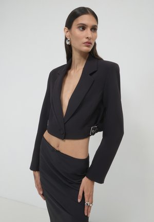 Cropped black blazer with peak lapels and side buckle detail, paired with a black high-waisted skirt, smooth fabric, and fitted silhouette.
