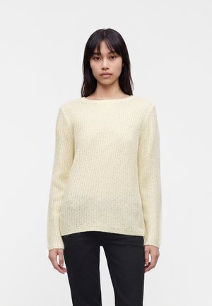 Jeune femme aux cheveux lisses et foncés portant un pull en tricot crème et un pantalon noir sur un fond blanc uni.