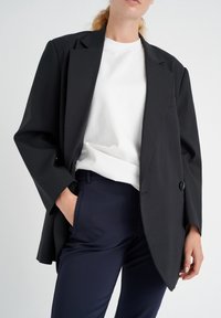 Černý oversized blazer s jedním knoflíkem, s širokými klopami. Nosený přes bílou košili a námořnické kalhoty. Hladká látka a uvolněný střih.