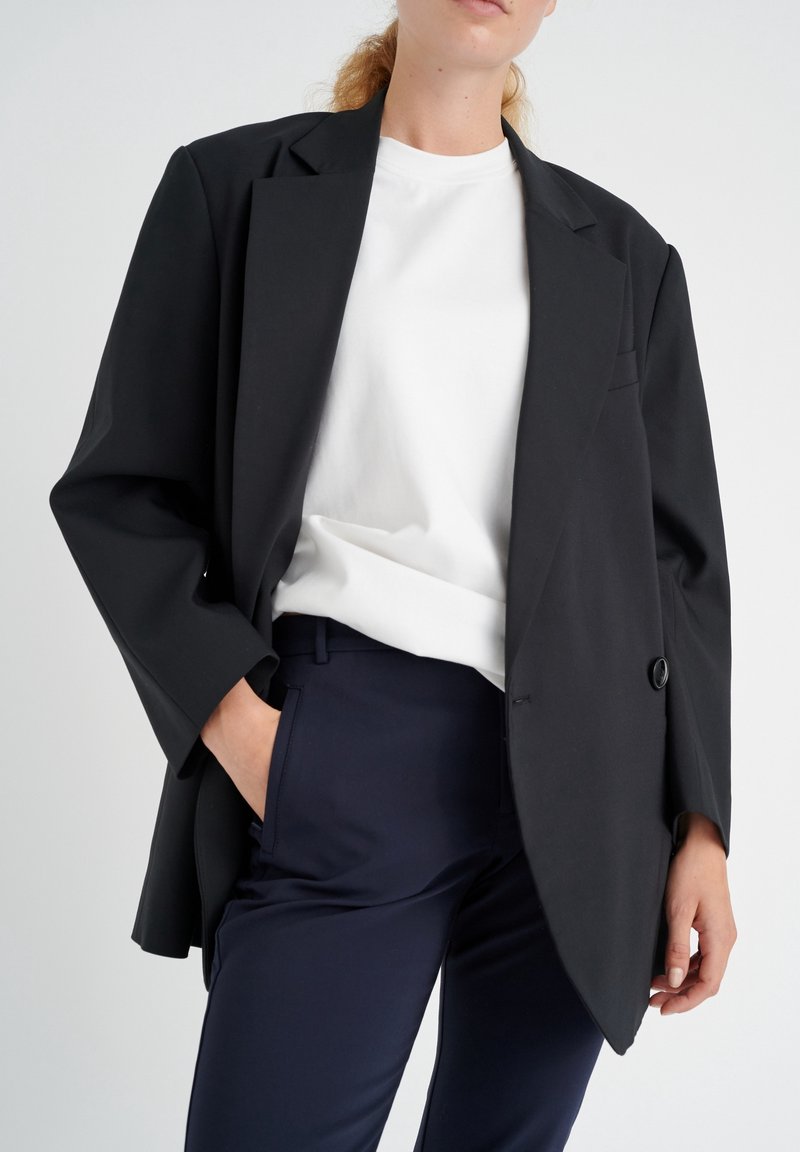 Černý oversized blazer s jedním knoflíkem, s širokými klopami. Nosený přes bílou košili a námořnické kalhoty. Hladká látka a uvolněný střih.