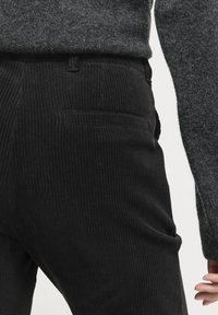 Svarta kordflöjelbyxor med vertikal ribbad textur, med en bakficka och en figurnära silhuett, ihop med en mörkgrå stickad tröja.