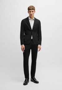 Zwart pak met een getailleerde blazer met twee voorkeizen en bijpassende getailleerde broek. Gecombineerd met een lichtgekleurde shirt en zwarte schoenen.