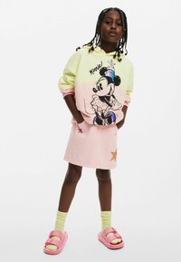 Felpa gialla pastello e rosa con grafica di Minnie Mouse, abbinata a una gonna di denim rosa con dettagli a stella. Sandali rosa e calzini gialli.