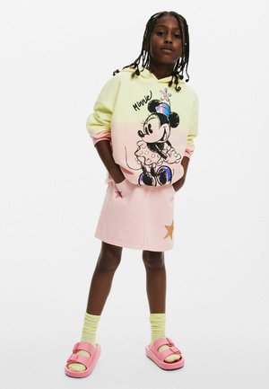 Pastellgelbe und rosa Kapuzenjacke mit Minnie Mouse Grafik, kombiniert mit einem rosa Jeansrock mit Sternen-Detail. Rosa Sandalen und gelbe Socken.