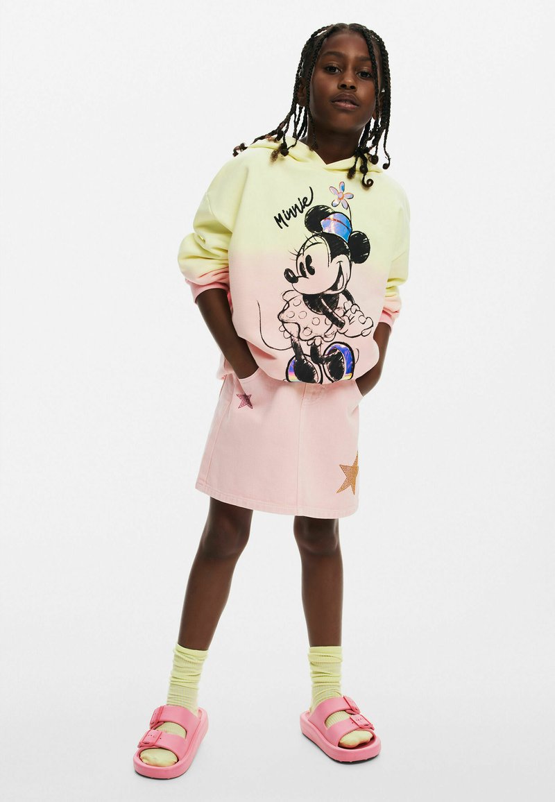 Felpa gialla pastello e rosa con grafica di Minnie Mouse, abbinata a una gonna di denim rosa con dettagli a stella. Sandali rosa e calzini gialli.