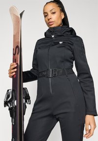 Μαύρη στολή σκι με εφαρμοστή σχεδίαση, μπροστινό φερμουάρ και ρυθμιζόμενη ζώνη. Μαύρα γυαλιά σκι και σκι με δεσίματα κρατημένα στο αριστερό χέρι.