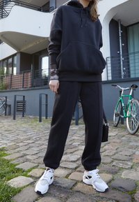 Černá fleecová mikina s kapucí a šňůrkou, v kombinaci s černými zúženými tepláky a bílými teniskami. Na pozadí se nachází moderní budova a bicykly.