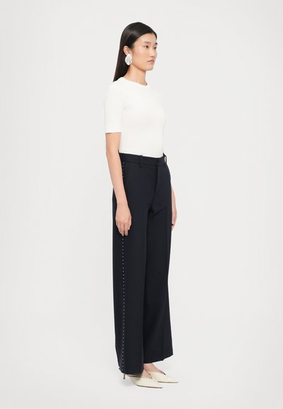 Wide-leg navy trousers with a tailored fit, featuring a side detail of silver studs and a smooth fabric texture. Paired with a white top and pointed shoes.