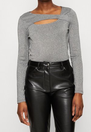 Mujer con una blusa gris de manga larga con un corte horizontal cerca del cuello y pantalones altos de cuero negro.