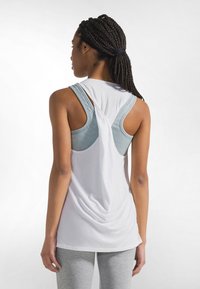 Valkoinen hihaton paita, jossa on laskostettu selkäosa, ja alla sininen ribbineuloksinen urheilurintaliivit. Malli käyttää harmaita legginssejä.