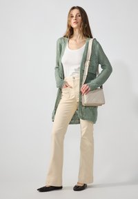 Απαλή πράσινη πλεκτή ζακέτα, λευκό αμάνικο μπλουζάκι, μπεζ παντελόνι ψηλής μέσης, μαύρες επίπεδες παπούτσια και μικρή μπεζ τσάντα ώμου.