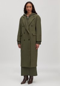 Olivgrüner, doppelt knöpfbarer Mantel mit Kapuze, großen Revers, seitlichen Taschen, kombiniert mit einem passenden Kleid und braunen spitzen Ankle Boots.